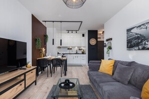 A modern living room with a sofa, coffee table, and TV. There is a dining area with chairs and a table. The kitchen is visible in the background.
