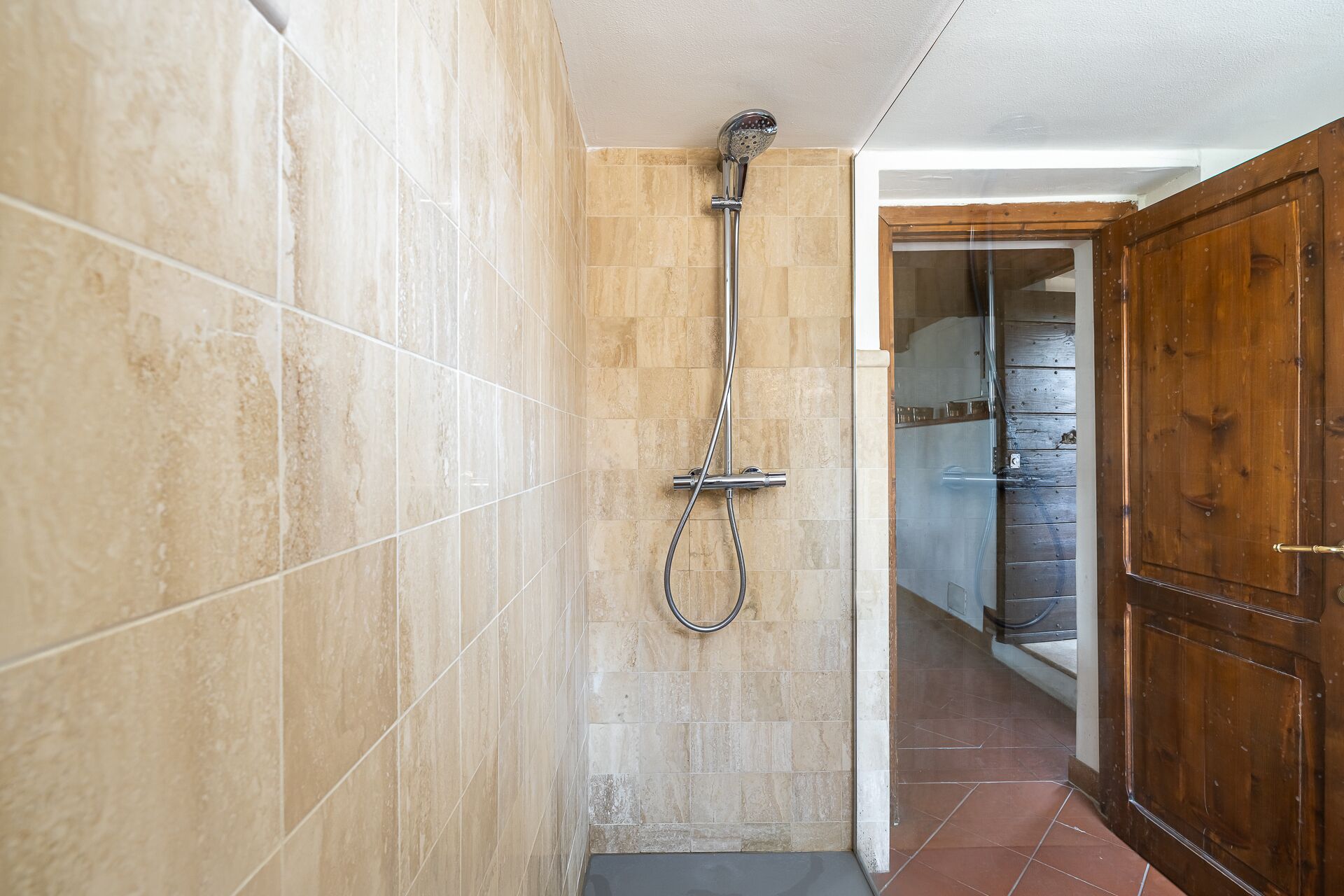 A shower with a handheld showerhead and a wall-mounted faucet, tiled walls, and a wooden door.