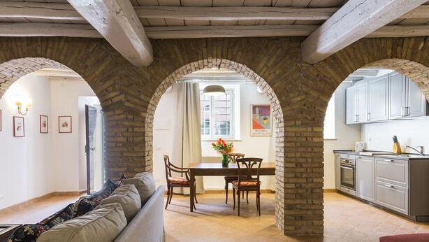 A dining area with a brick archway, a wooden table, and chairs. There is a kitchen area with cabinets and a sink visible through the archway. - Castello del Duca (Rome)