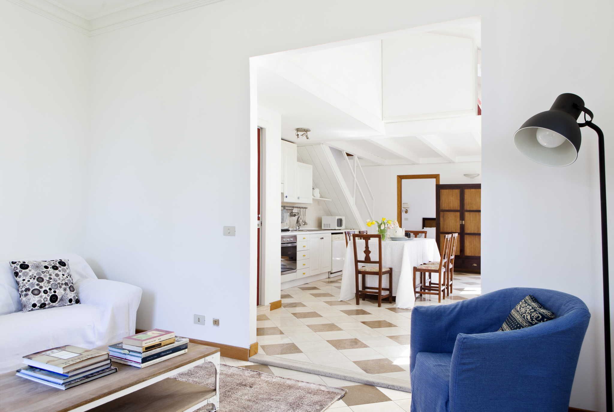 A modern living room with a white sofa, a blue armchair, and a coffee table with books. A kitchen is visible in the background with white cabinets and a dining area.