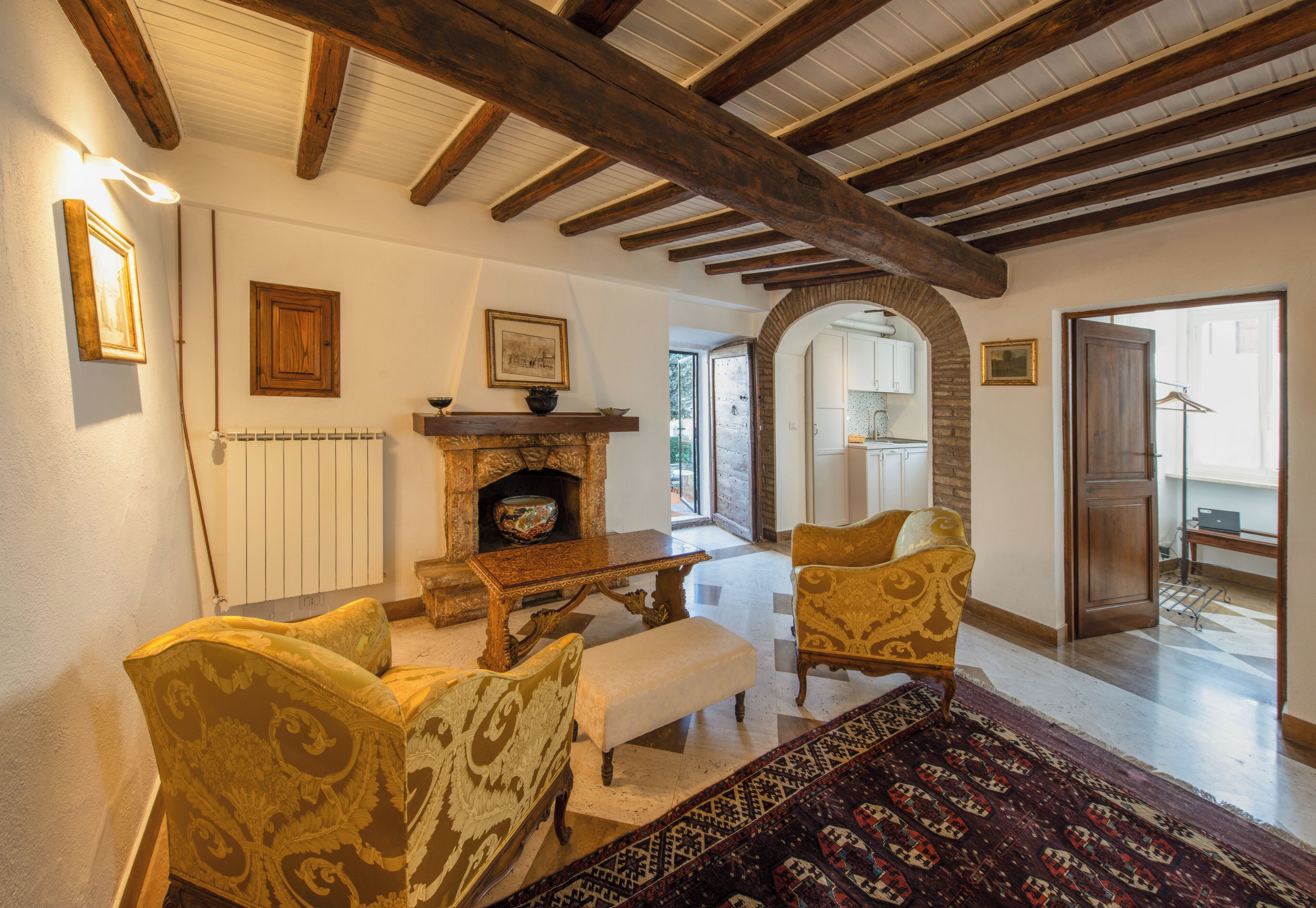 A traditional living room with wooden beams, a fireplace, patterned armchairs, and a view of the kitchen through an archway.