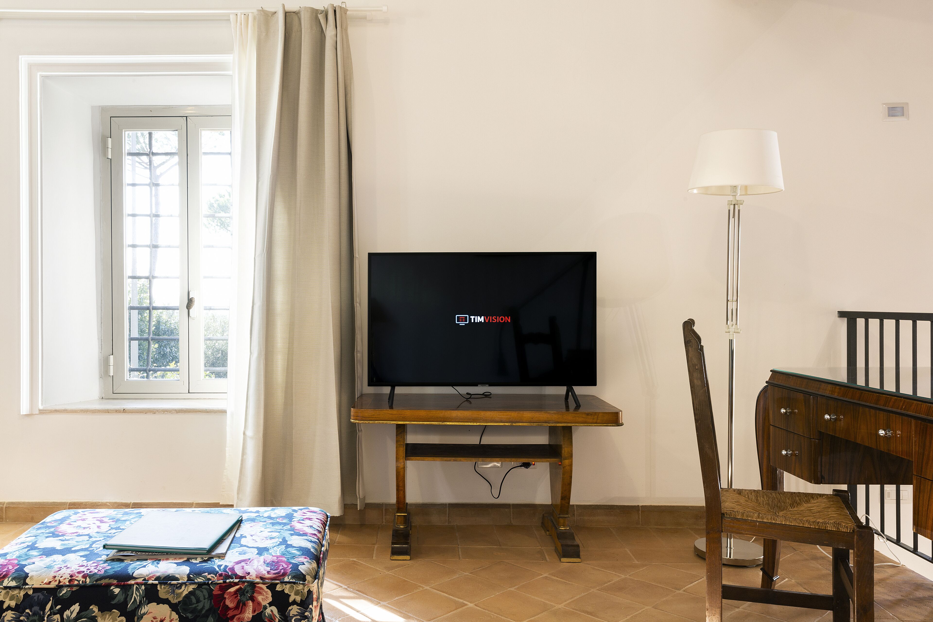 Una habitación con una televisión sobre un mueble de madera, una silla, una mesita con un cuaderno y una ventana con cortinas.