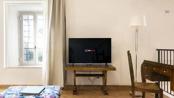 Una habitación con una televisión sobre un mueble de madera, una silla, una mesita con un cuaderno y una ventana con cortinas.