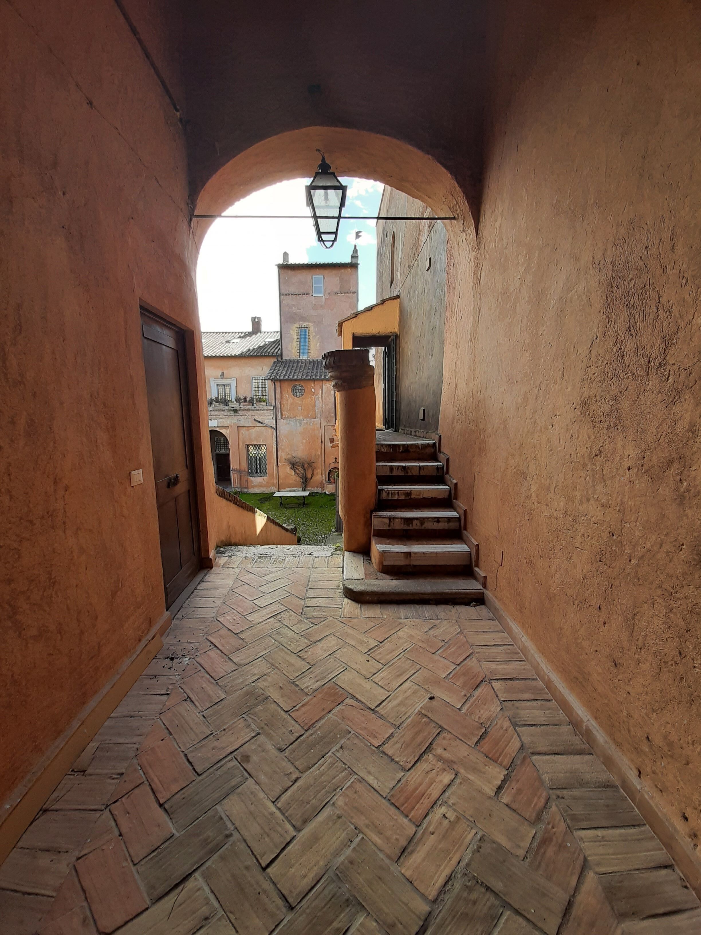 A narrow alleyway with a herringbone-patterned brick floor, a wooden staircase, and a stone archway.