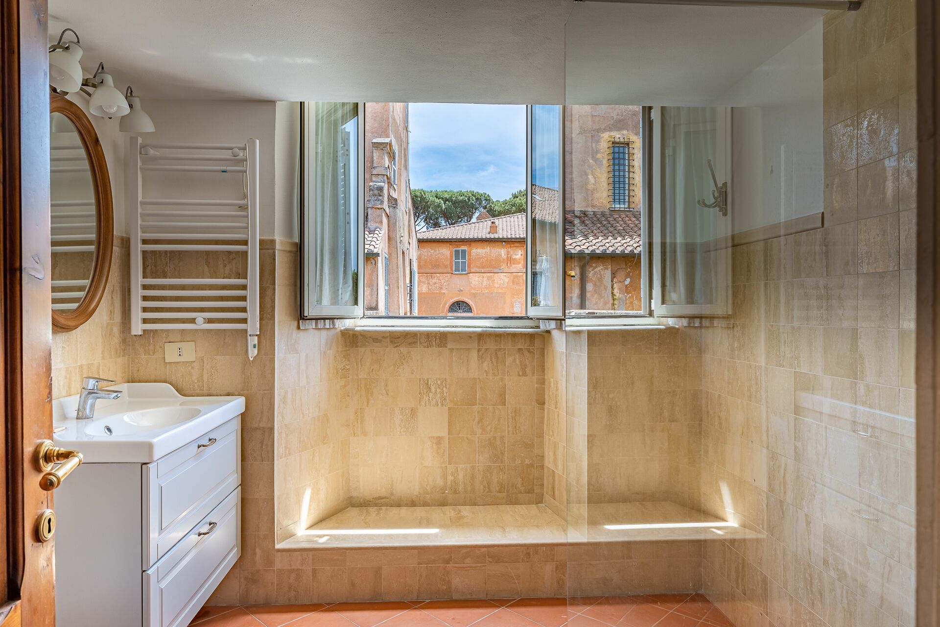 A bathroom with a bathtub, a sink, and a mirror. There is a window with a view of buildings outside.