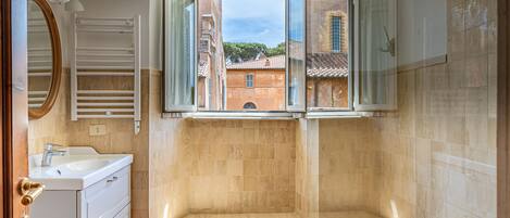 A bathroom with a bathtub, a sink, and a mirror. There is a window with a view of buildings outside.