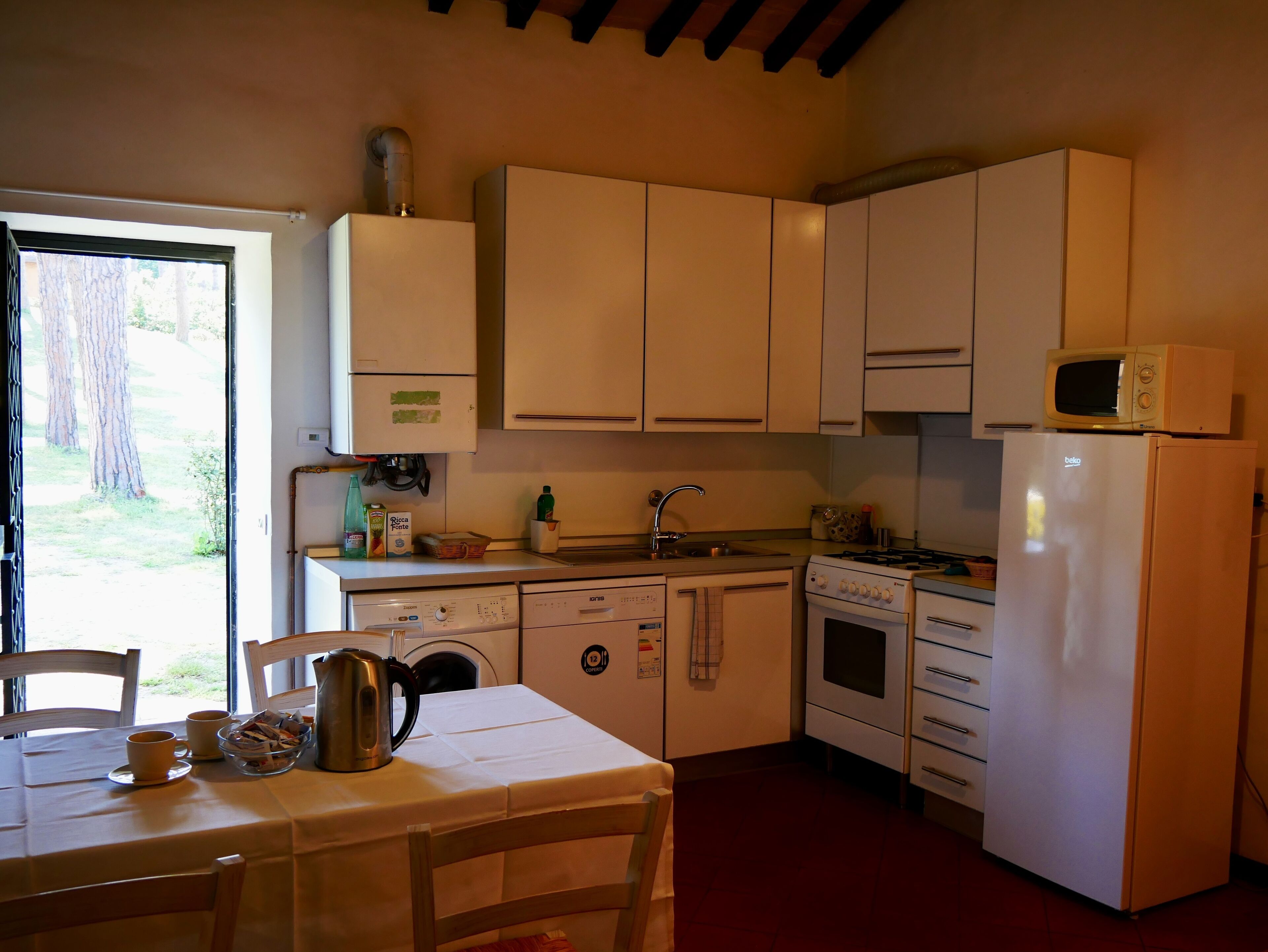 Cocina compacta con gabinetes blancos, microondas, estufa y pileta. Hay una mesa con sillas y una ventana que proporciona luz natural.