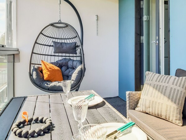 A balcony with a hanging chair, a table with a glass, a plate, and a bracelet.
