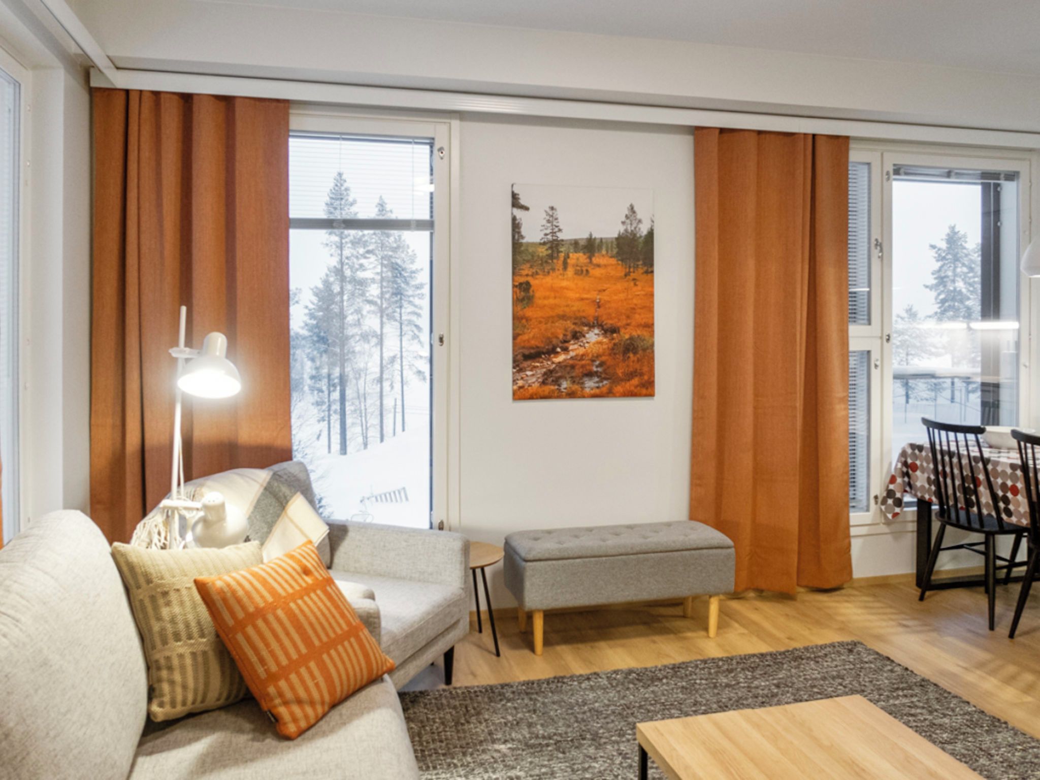 A living room with a sofa, a table, a bench, a lamp, a painting, and a dining area with chairs.