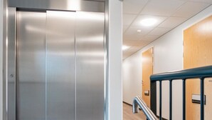 A modern elevator with a metallic finish, a wooden door, and a metal handrail in a hallway with tiled flooring.