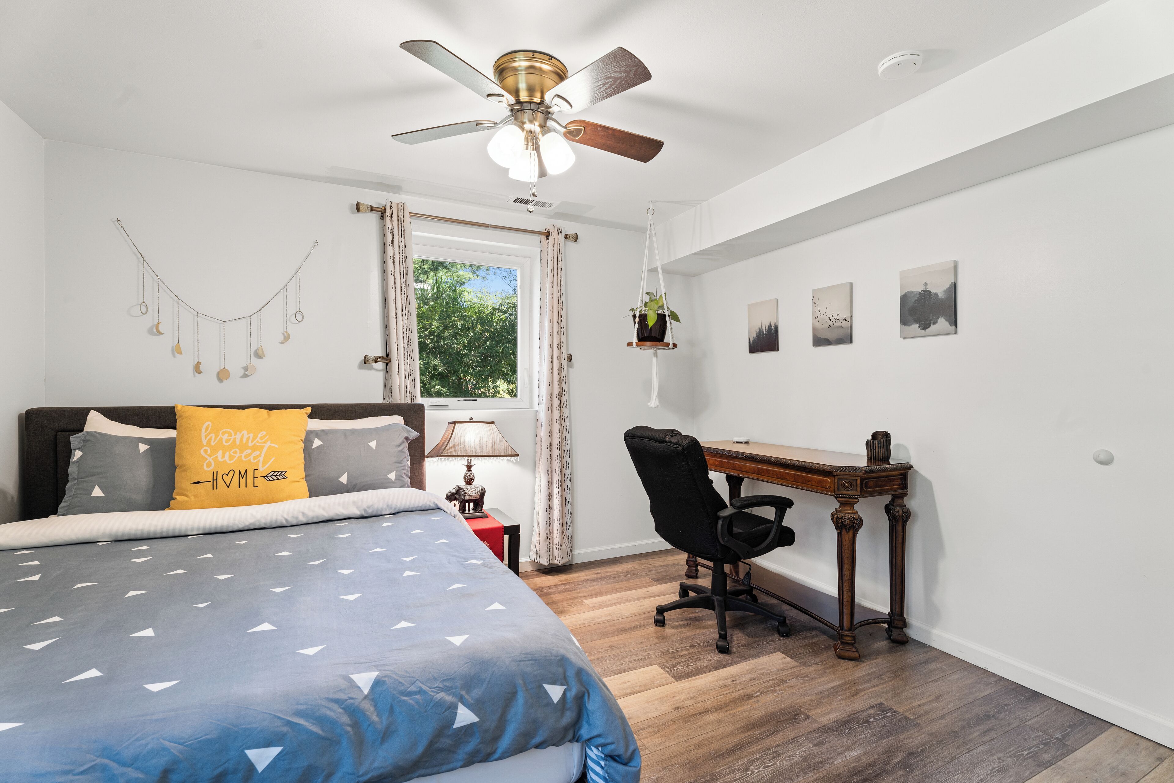 A bedroom with a bed, a desk, a chair, a ceiling fan, and a window.