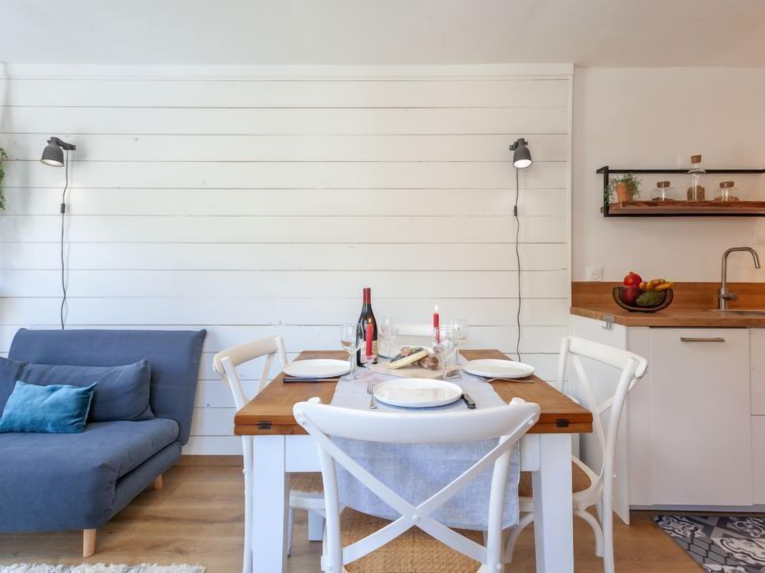 Un coin repas avec une table en bois, des chaises blanches et un canapé bleu. Deux lampes suspendues sont placées au-dessus de la table, et un espace cuisine comprend une étagère où sont rangés des bocaux et un saladier rempli de fruits.