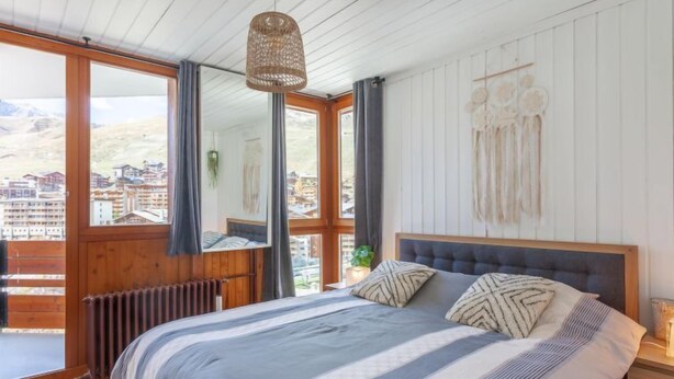 A bedroom with a large bed, wooden floor, a view of buildings through a window, and a hanging light fixture. - Savoy (Lavachet) by Interhome (Tignes)