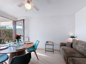 A dining area with a round table, chairs, and a vase of flowers. There is a ceiling fan above and a sofa in the background.
