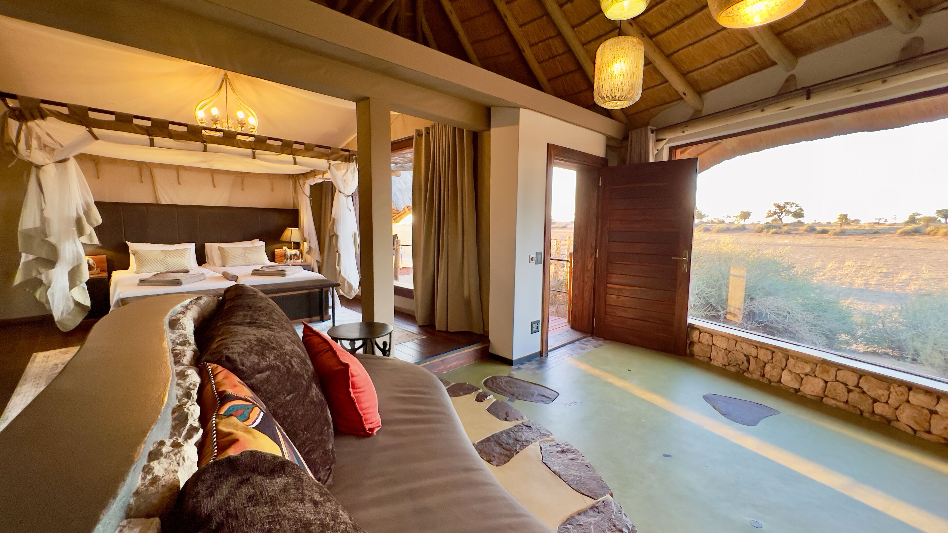 Un dormitorio con cama, sofá, candelabro, ventana con vista al campo y puerta de madera.