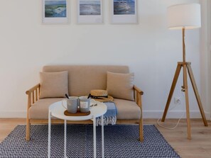 Uma sala de estar aconchegante com um sofá bege, uma mesa redonda pequena com duas canecas, um abajur e três quadros na parede.