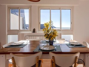 Uma sala de jantar com uma mesa de madeira posta para dois, duas cadeiras, um vaso com flores amarelas e vista para o mar através de grandes janelas.