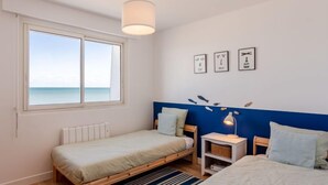 Ein Schlafzimmer mit zwei Betten, einem Fenster mit Meerblick, einem weißen Heizkörper, einem blauen Kopfteil, einem weißen Nachttisch und einer Lampe.