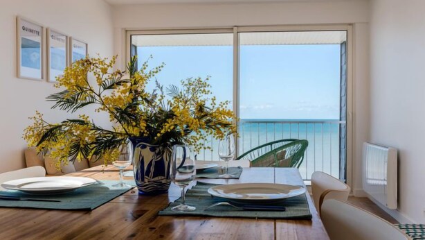 Ein Essbereich mit einem Holztisch, gedeckt für zwei Personen, einer Vase mit gelben Blumen und Meerblick durch große Fenster.