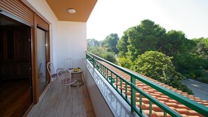Un balcon doté d’un plancher en bois, de deux chaises, d’une petite table sur laquelle sont disposés des fruits, et qui offre une vue sur des arbres verdoyants et un toit recouvert de tuiles rouges.