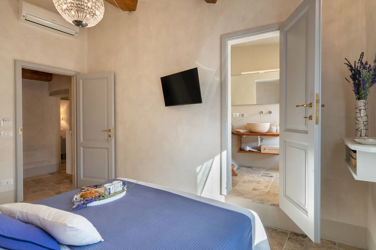 Un dormitorio con cama, televisor de pantalla plana, baño con lavamanos y espejo, y una puerta que da a otra habitación.