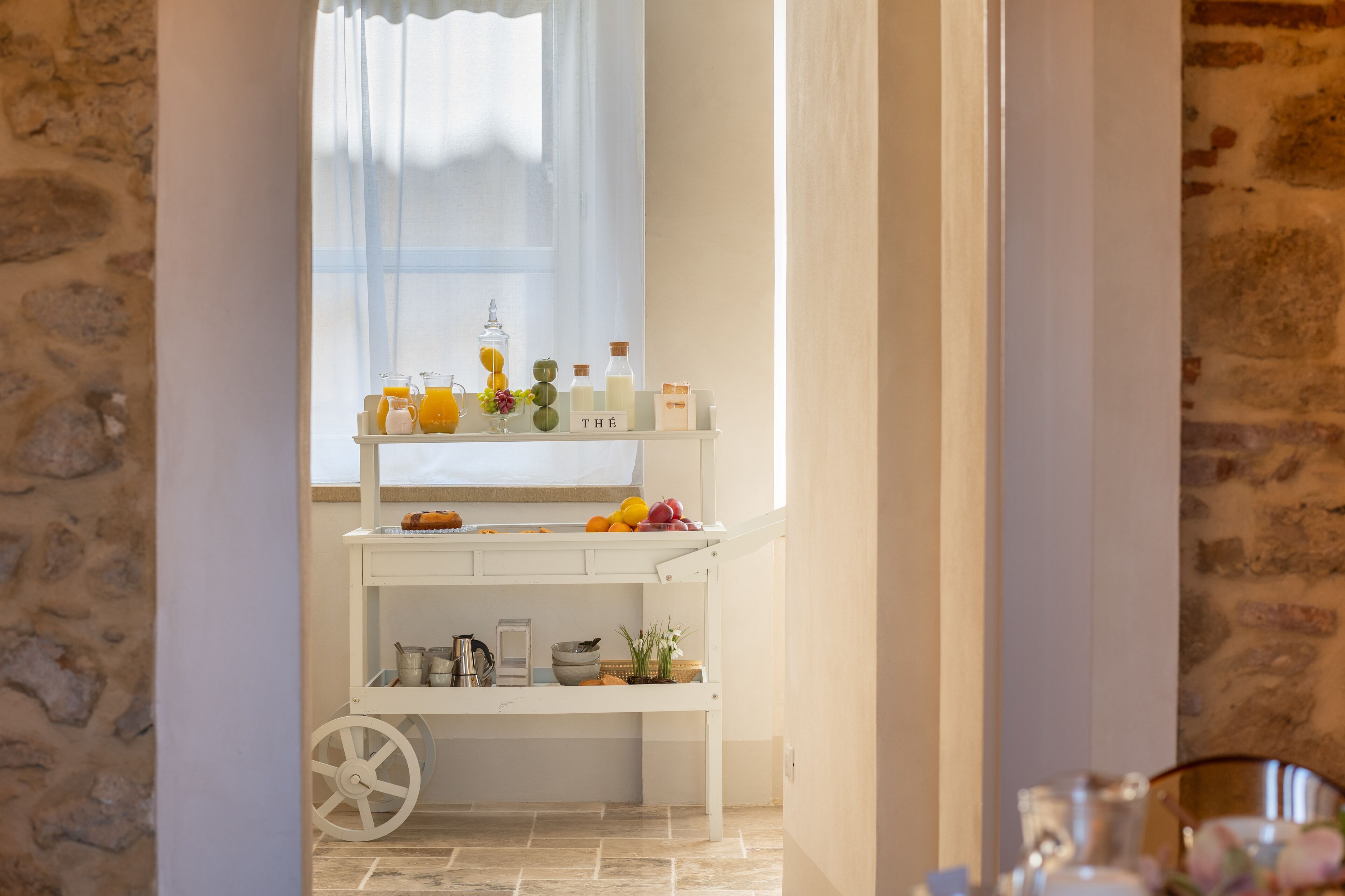 Una zona de cocina con un carrito blanco, estantes con utensilios de cocina y una ventana con cortinas traslúcidas.