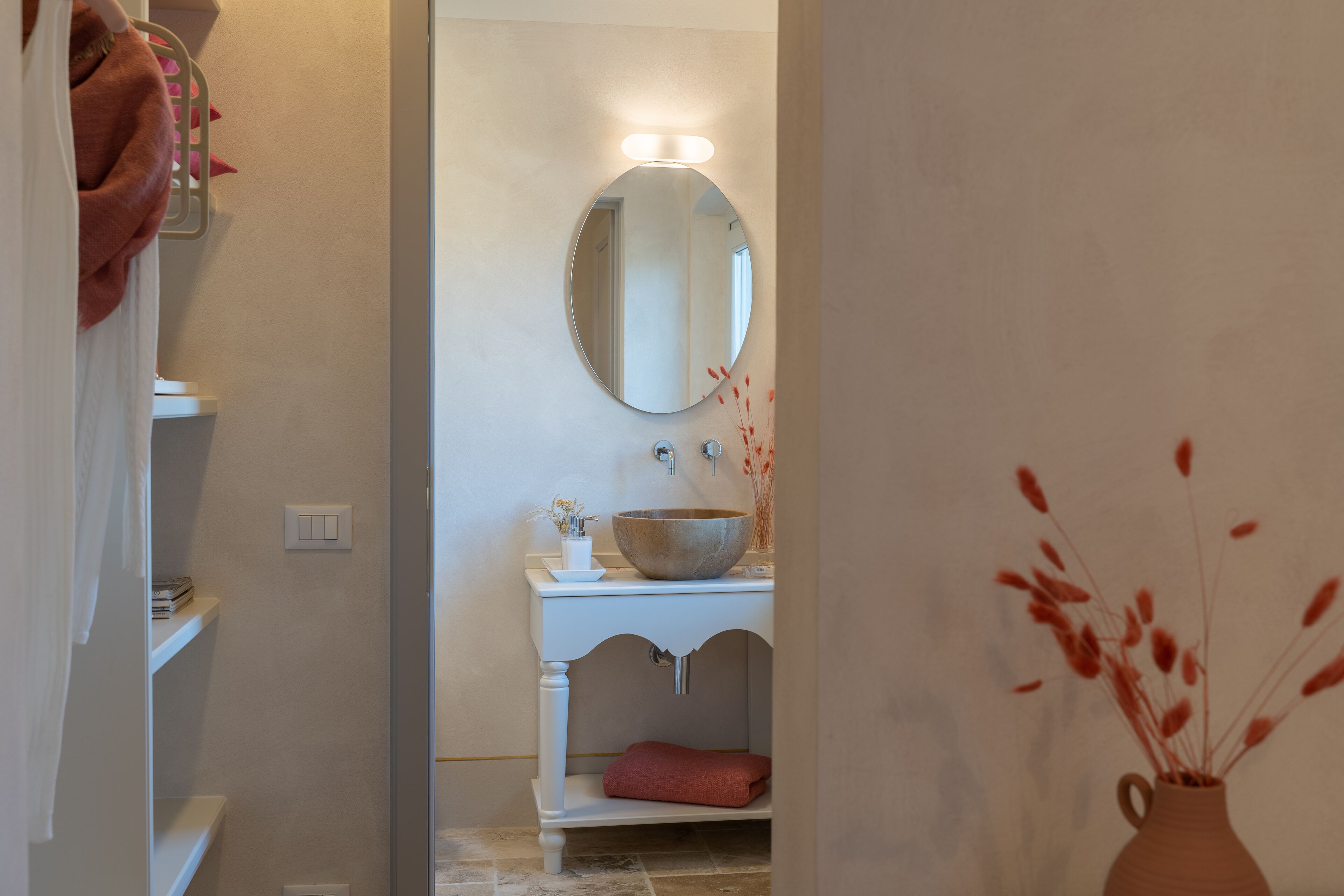 A bathroom with a round mirror, a white sink, and a vase with dried plants.