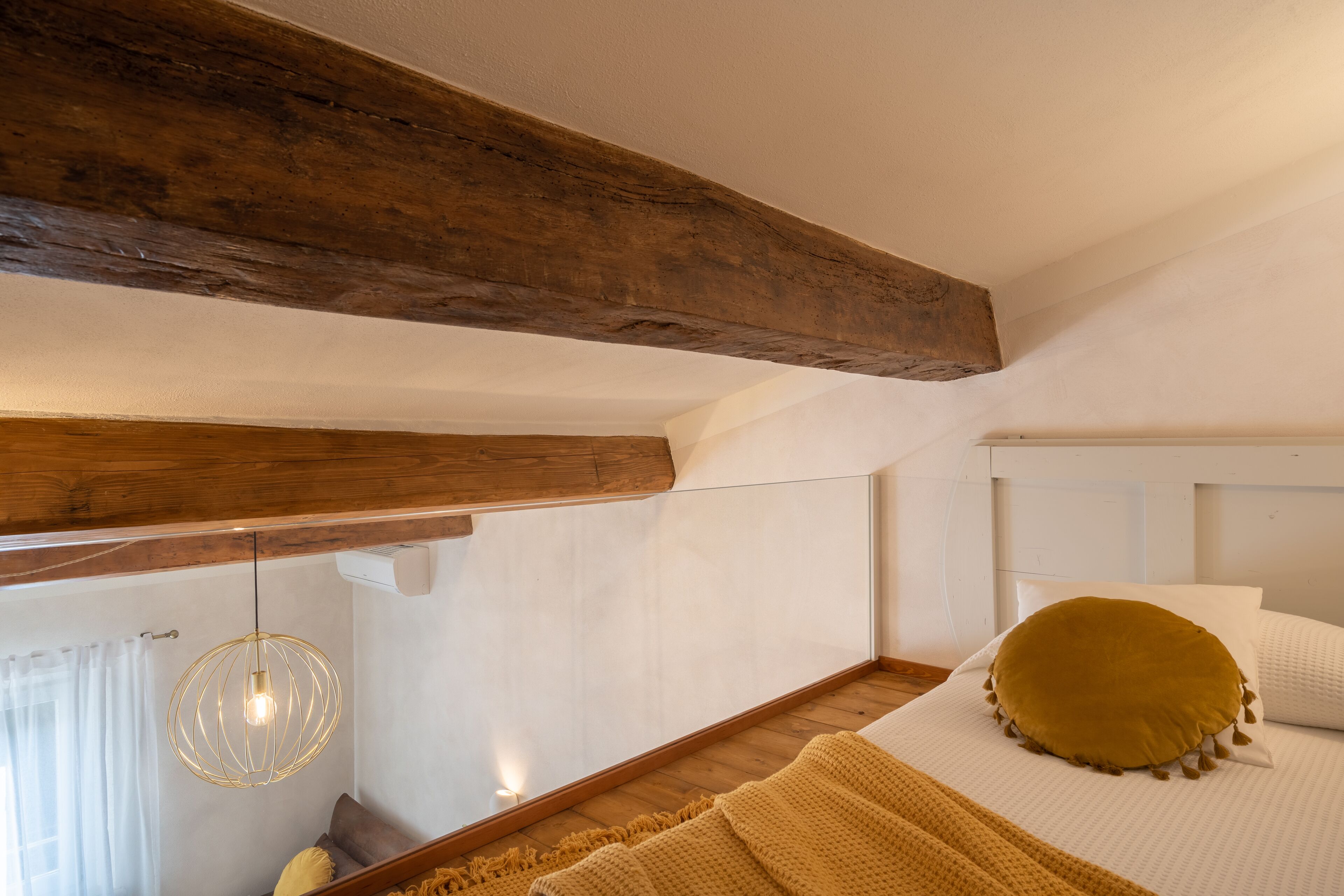 Une chambre avec des poutres apparentes, un lit avec un oreiller jaune, un luminaire suspendu et une fenêtre avec des rideaux.