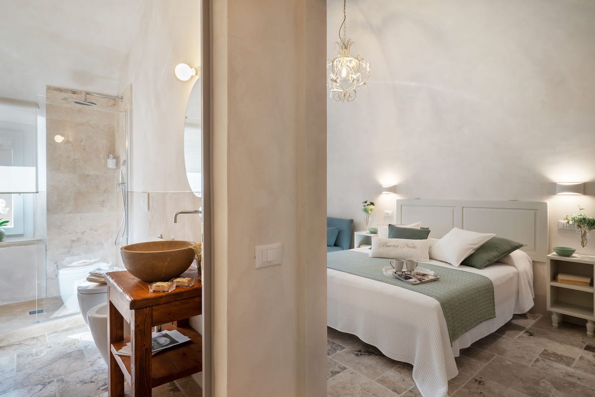 Un baño moderno con un lavamanos grande, un espejo y una ducha. El dormitorio tiene una cama con una manta verde, una mesita de noche y una mesita con una lámpara.