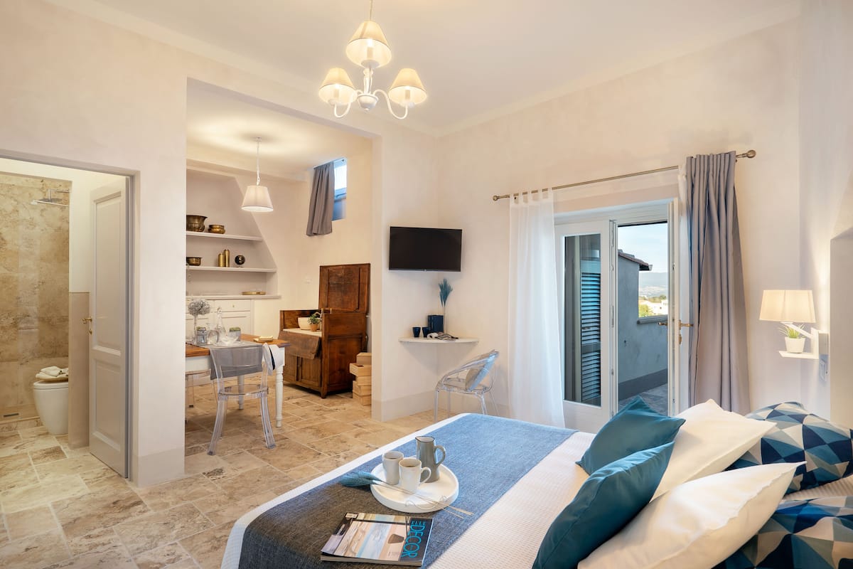 Una habitación de hotel moderna con una cama, zona de comedor, televisión y balcón con cortinas.