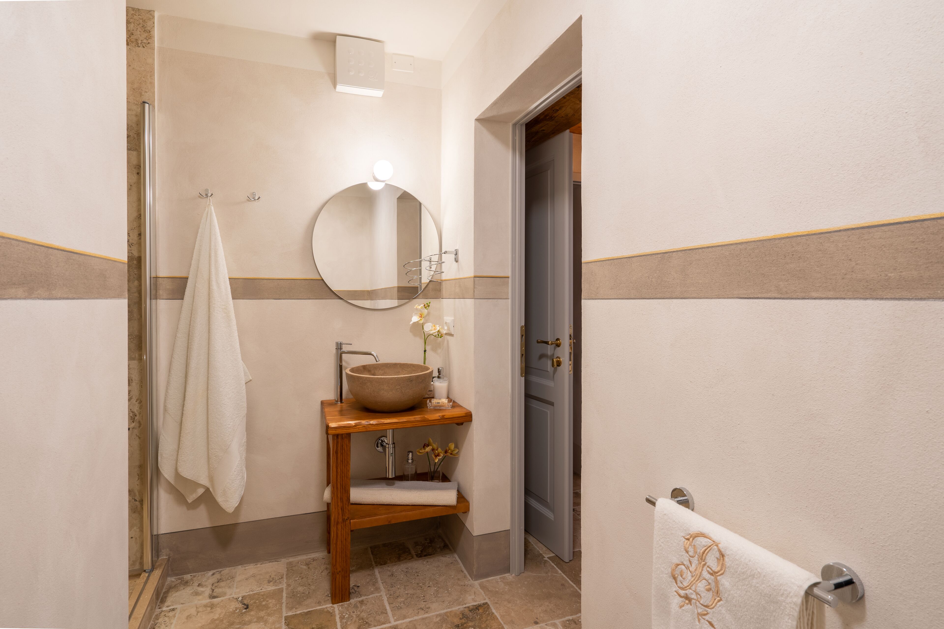 A bathroom with a wooden vanity, a round mirror, a large vessel sink, and a towel rack.