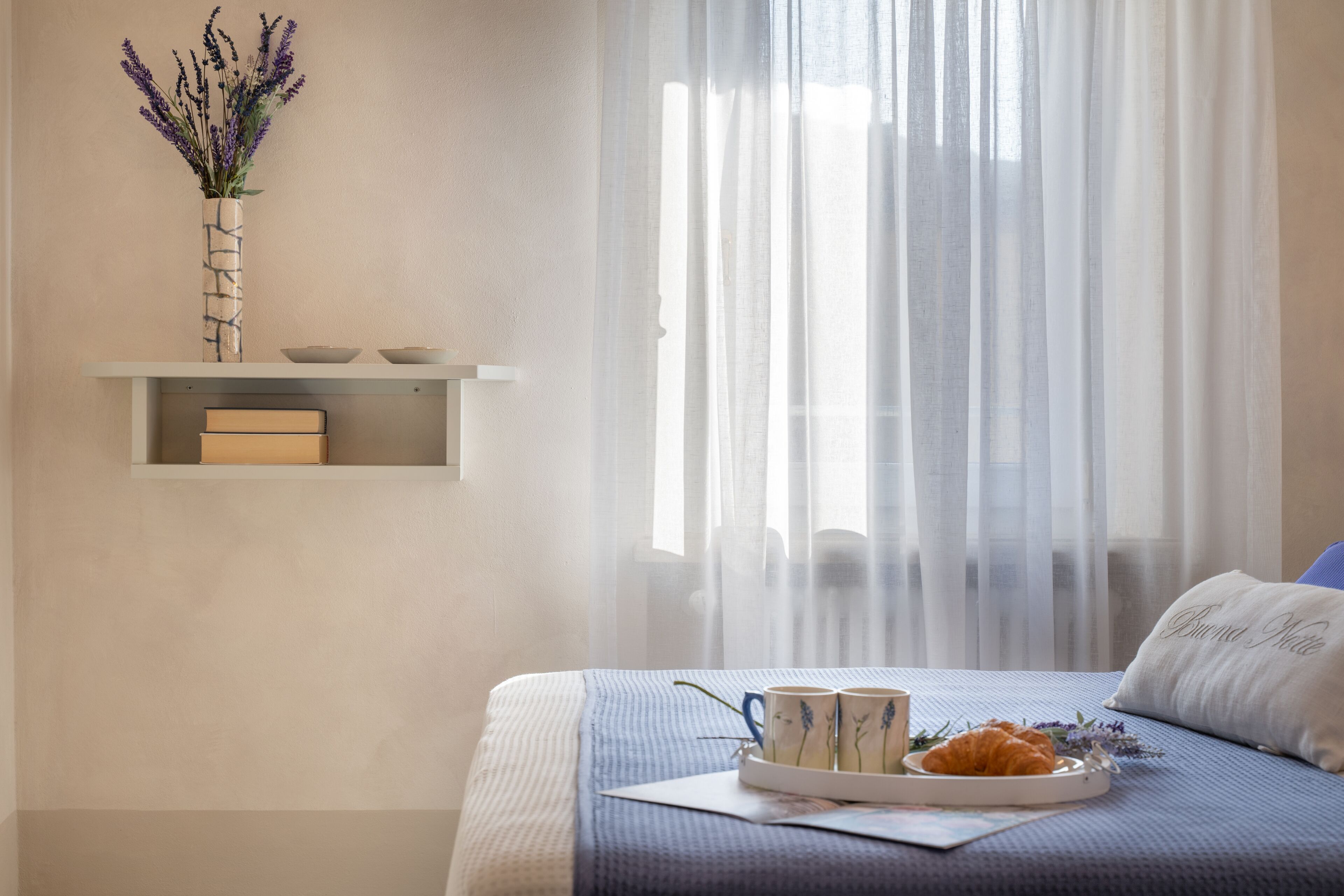 Un dormitorio ordenado con cama, mesita de noche con una bandeja de croissants y tazas de café, un jarrón con flores en una estantería y cortinas traslúcidas.