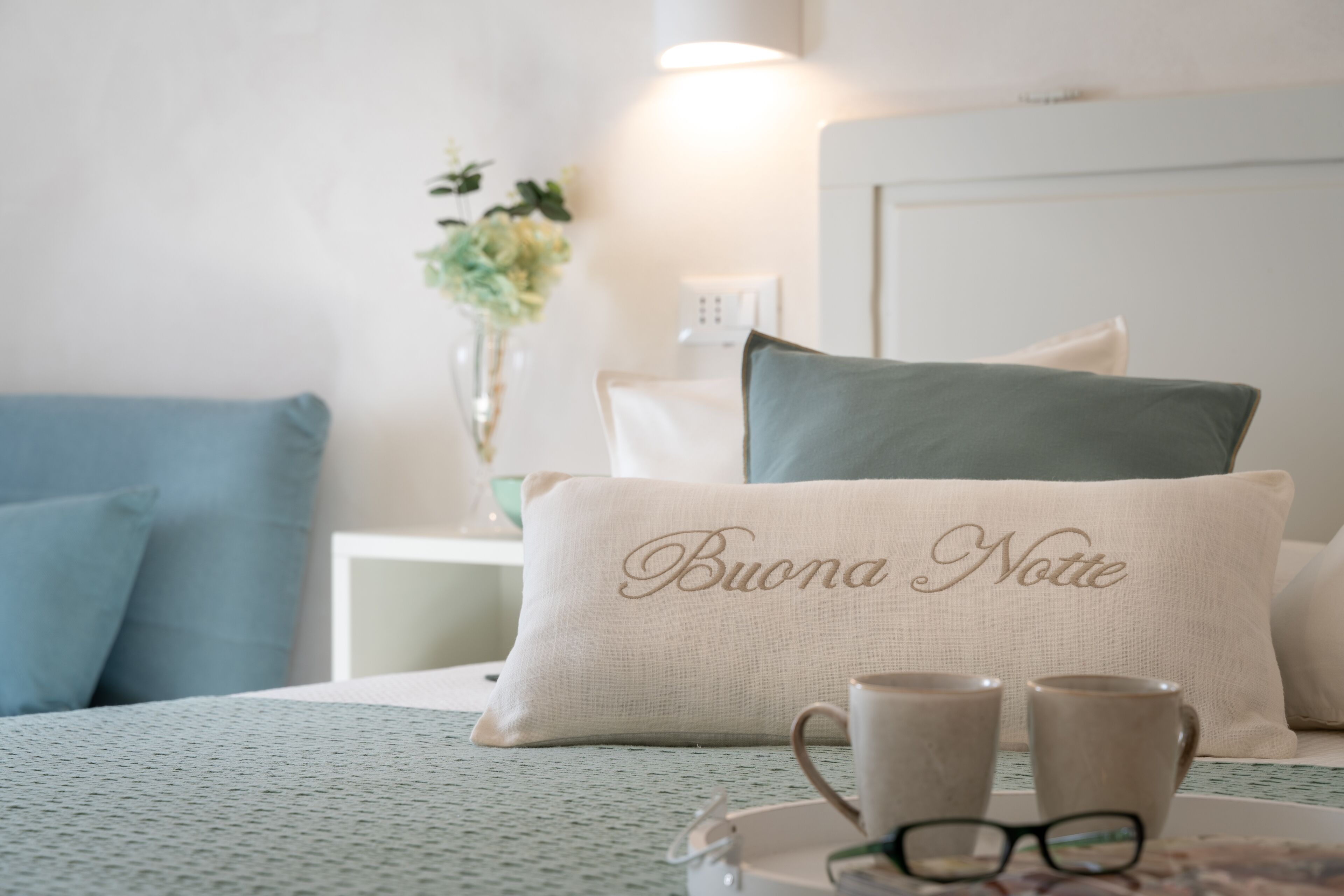 A neatly arranged bedroom with a bed, pillows, a bedside table with a lamp, and a vase with flowers.