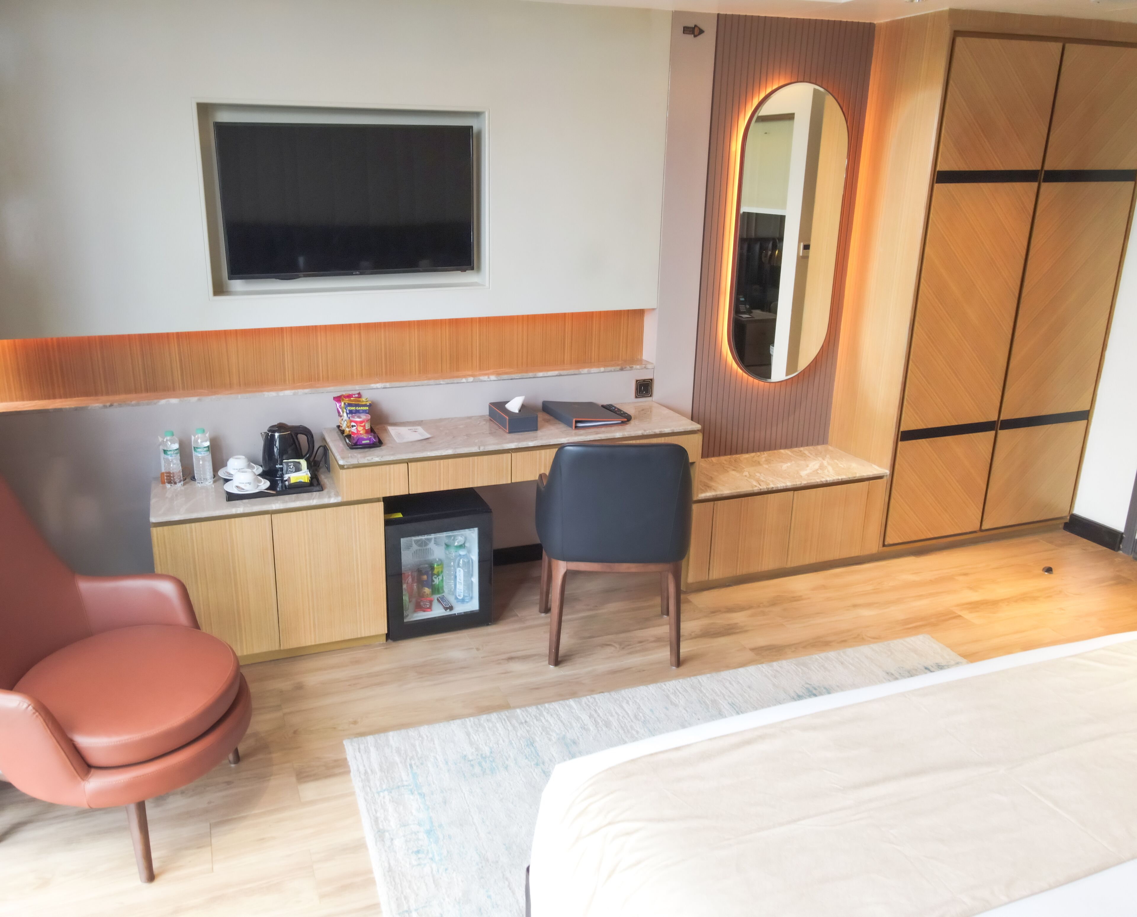 Una habitación de hotel con cama, escritorio con silla, refrigerador pequeño, espejo y ventana.