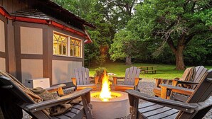 A cozy outdoor seating area with Adirondack chairs around a central fire pit, set against a backdrop of trees and a wooden building. - Multi-Family Haven! 22ppl, Hot Tub, Golf (Pleasant View)