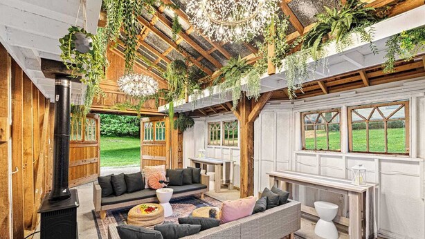 An open-plan living area with a fireplace, wicker furniture, hanging plants, and large windows overlooking a garden. - Multi-Family Haven! 22ppl, Hot Tub, Golf (Pleasant View)