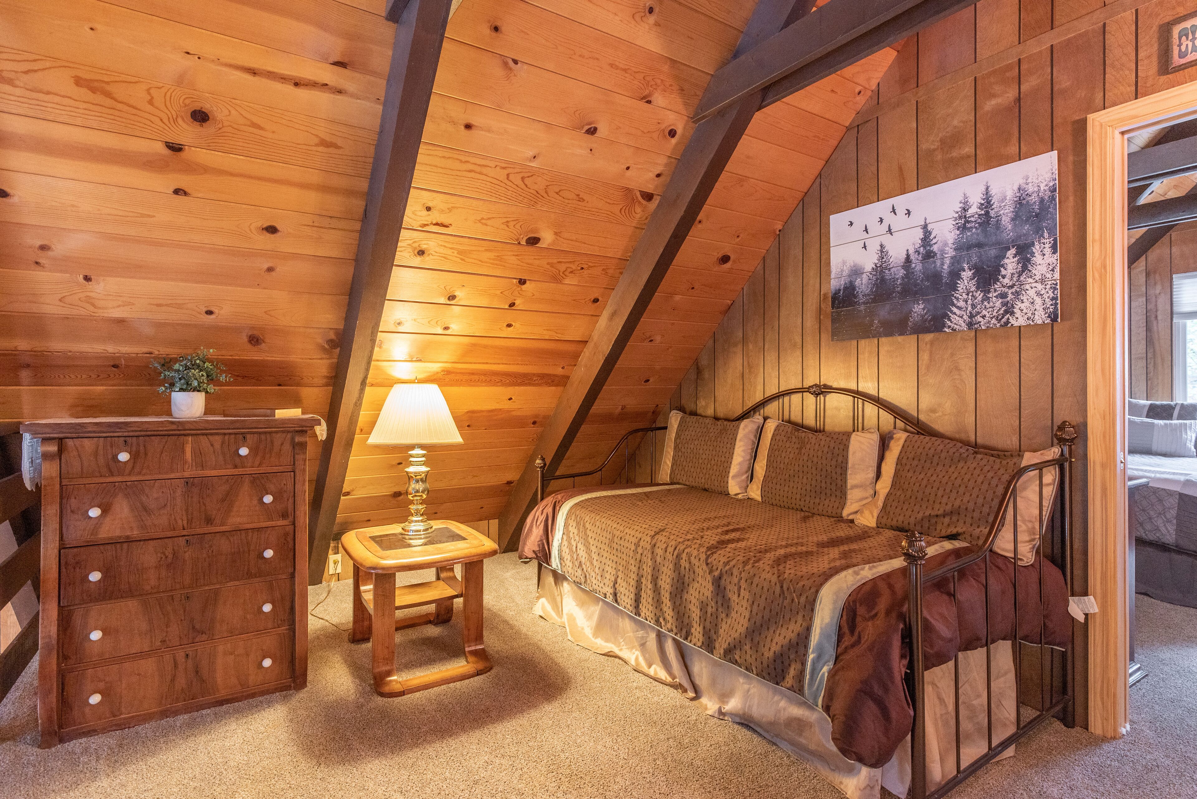 A cozy bedroom with a wooden ceiling, a bed with a patterned bedspread, a dresser, and a small table with a lamp.