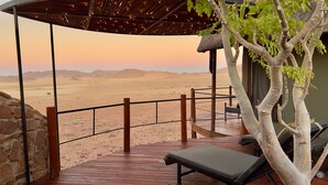 A desert view from a covered outdoor area with a wooden deck, lounge chairs, and a thatched roof structure. - Namib Outpost l Ondili (Sesriem)