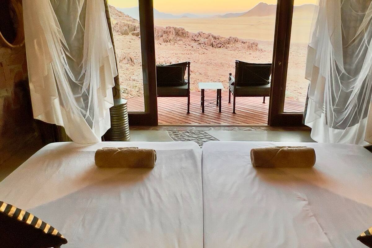 A room with two beds, a view of a desert landscape, and a wooden deck outside.