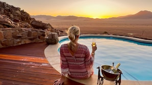 A person relaxing by a pool with a sunset view, a stone wall, and a deer's head decoration. - Namib Outpost l Ondili (Sesriem)