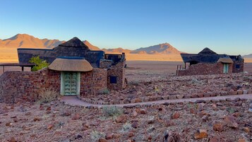Paisaje desértico con edificios de piedra y techos de paja, con una vista despejada de montañas lejanas.