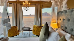 A bedroom with a large bed, a small table, two chairs, and a view of a desert landscape. - Namib Outpost l Ondili (Sesriem)