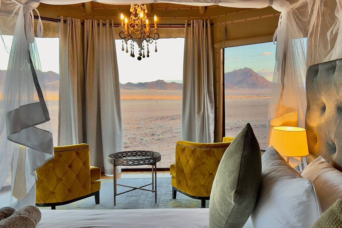 A bedroom with a large bed, a small table, two chairs, and a view of a desert landscape.