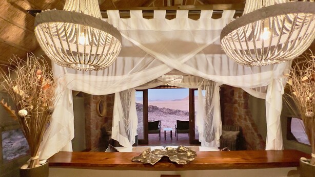 A room with a wooden reception desk, a view of a desert landscape through an open doorway, and a chandelier with a woven design. - Namib Outpost l Ondili (Sesriem)