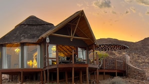 A wooden cabin with a thatched roof, large windows, and a balcony overlooking a rocky landscape. - Namib Outpost l Ondili (Sesriem)