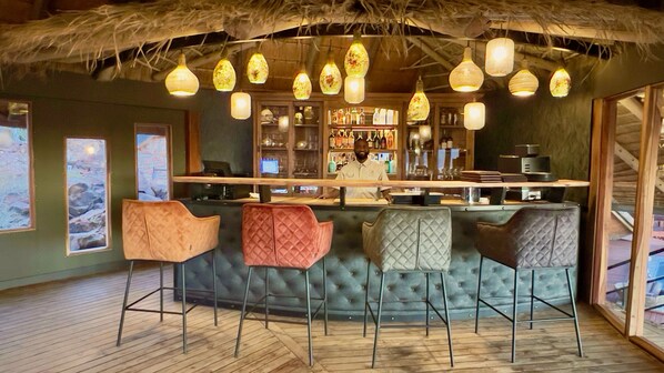 A bar area with a wooden bar counter, multiple stools, hanging pendant lights, and a rustic ceiling. - Namib Outpost l Ondili (Sesriem)