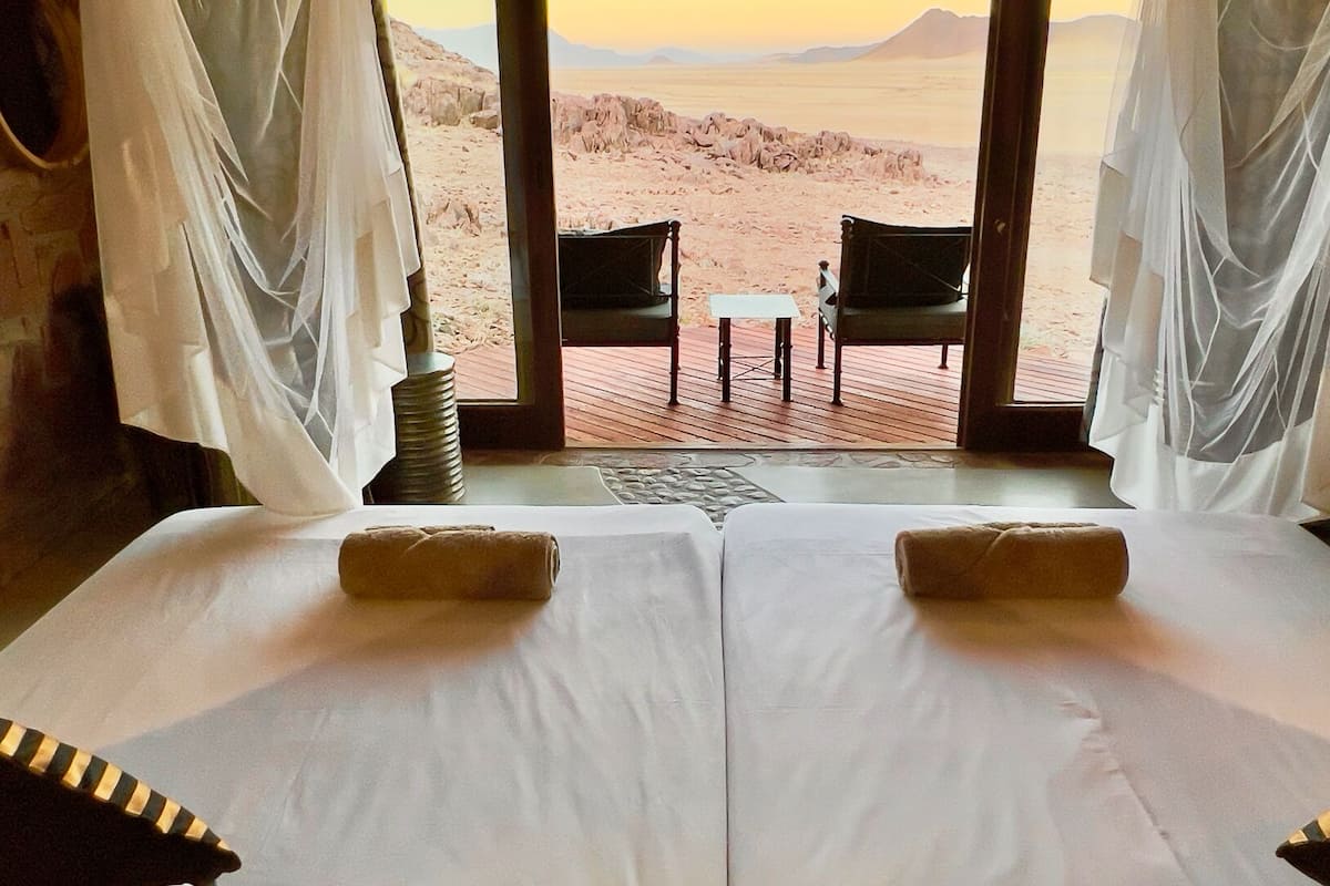 A room with two beds, a view of a desert landscape, and a wooden deck with chairs.