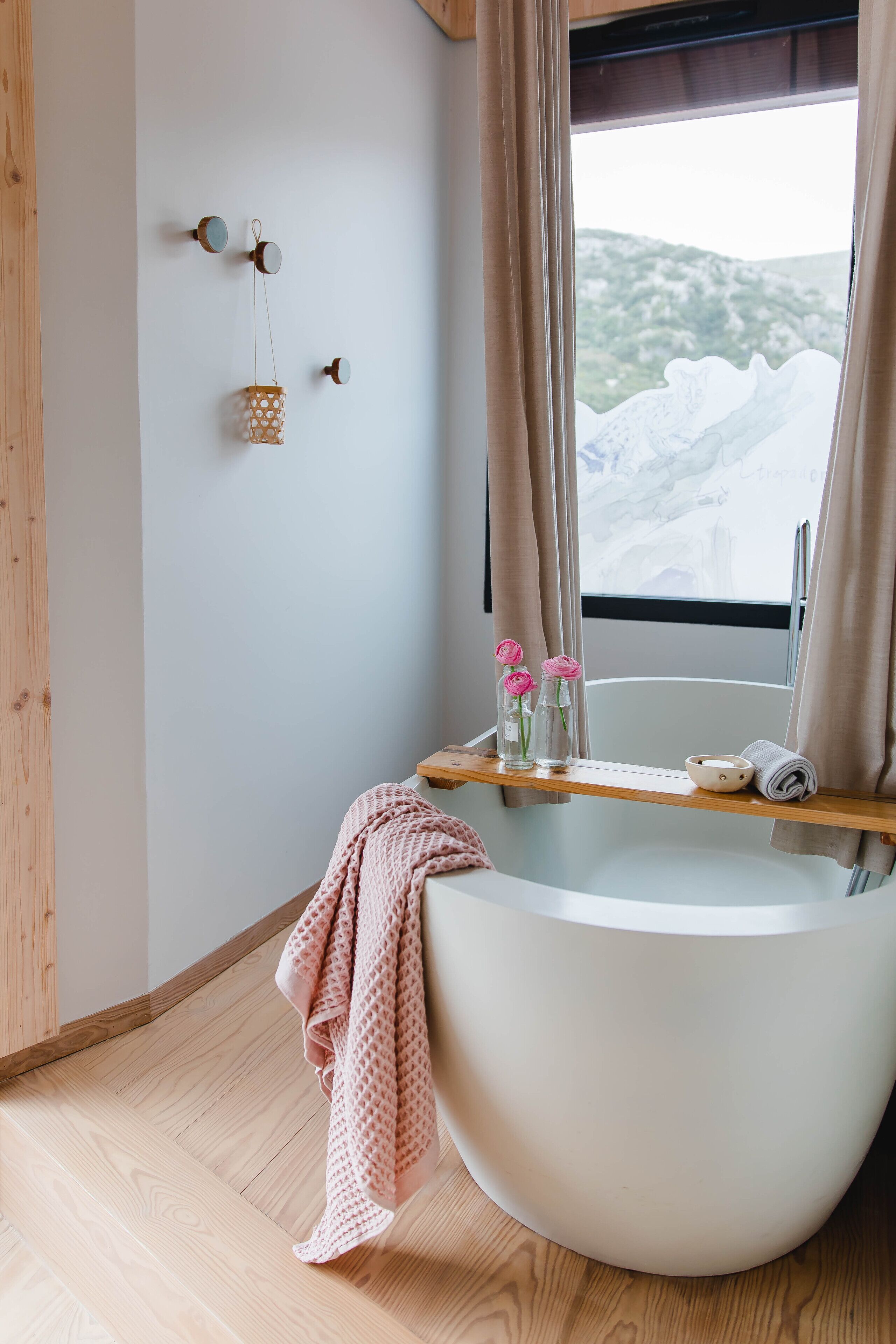 A modern bathroom with a freestanding bathtub, wooden flooring, a window with a view of a mountainous landscape, and a shelf with toiletries.