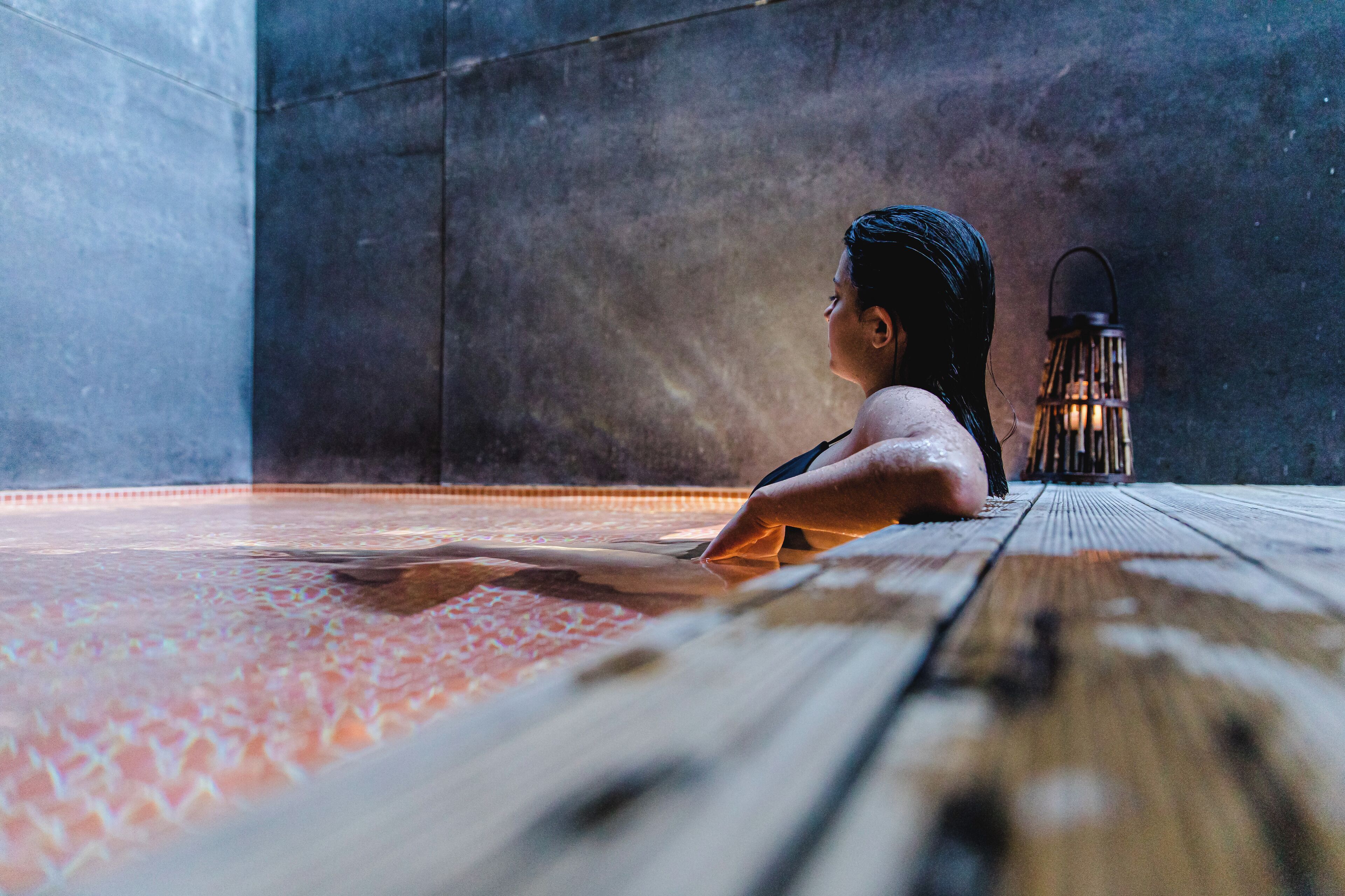 A person sitting on a wooden floor with a patterned rug, a lantern, and a textured wall in the background.