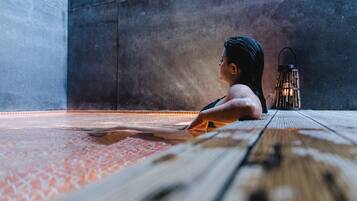 A person sitting on a wooden floor with a patterned rug, a lantern, and a textured wall in the background.